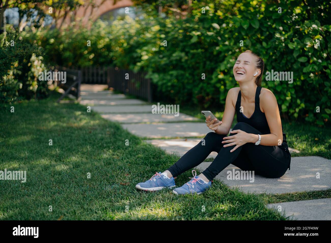 Overjoyed sportswoman sits on path laughs out uses smartphone for listening music from playlist share content and chatting dressed in active wear Stock Photo