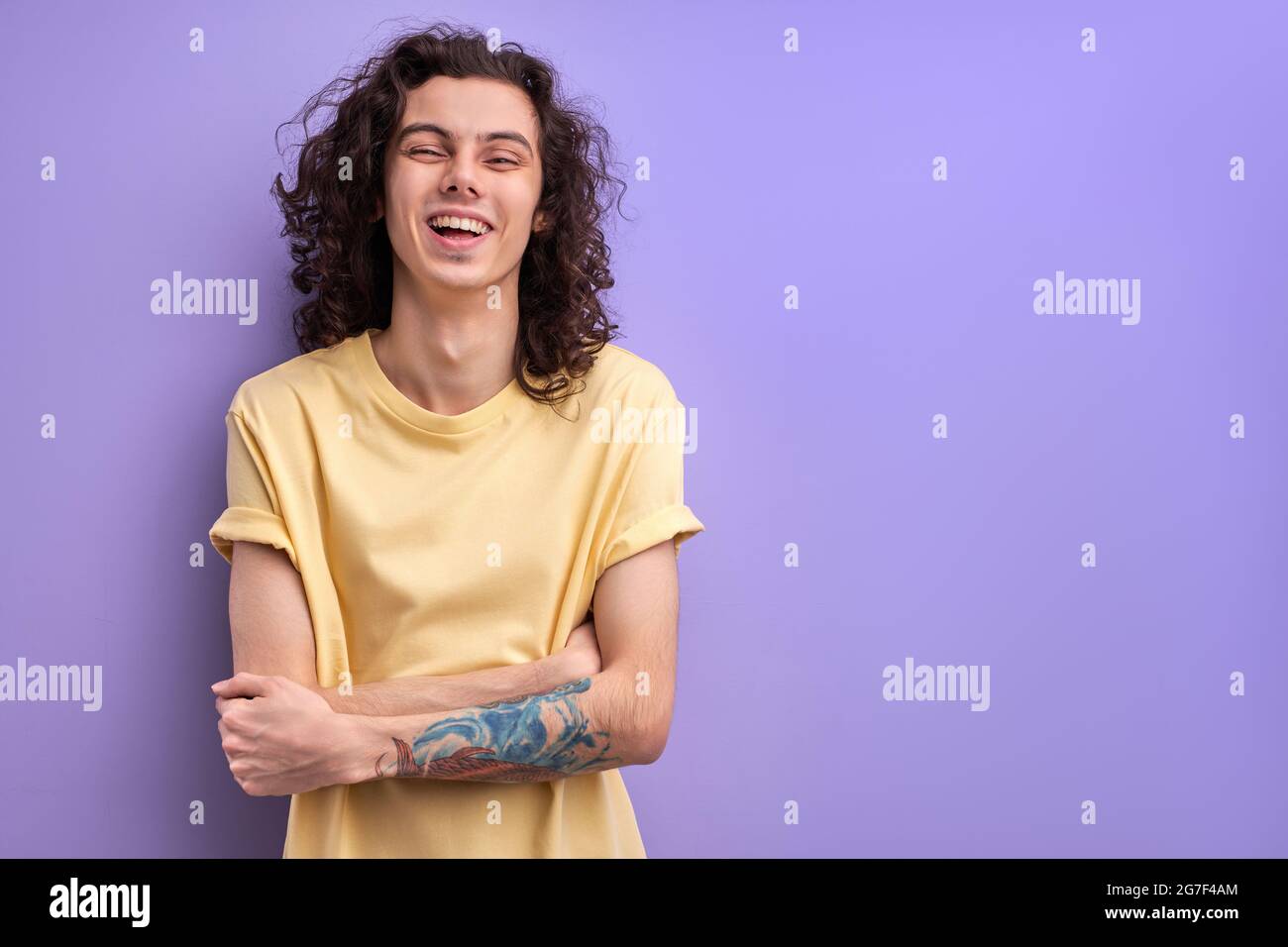 Positive Brunette Guy With Long Curly Hair Laughing, Standing With Arms Folded. Man Is In Good Mood, Happy Guy In Casual Wear Enjoys Life Stock Photo