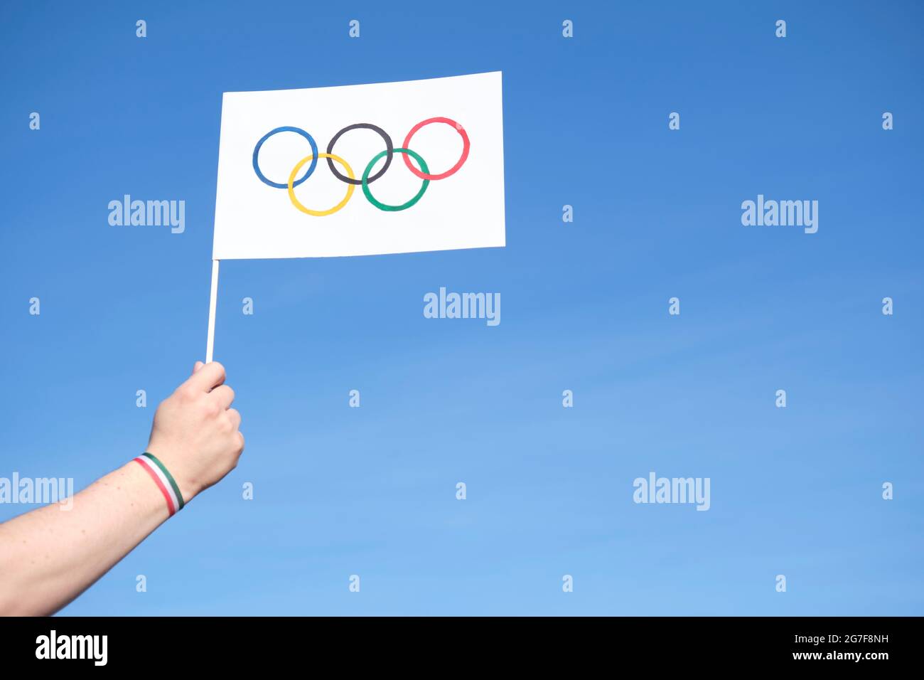 Hand holding a handmade Olympic Games flag outdoors, against a clear sky, wearing a green, white and red bracelet, colors of Mexico and Italy national Stock Photo