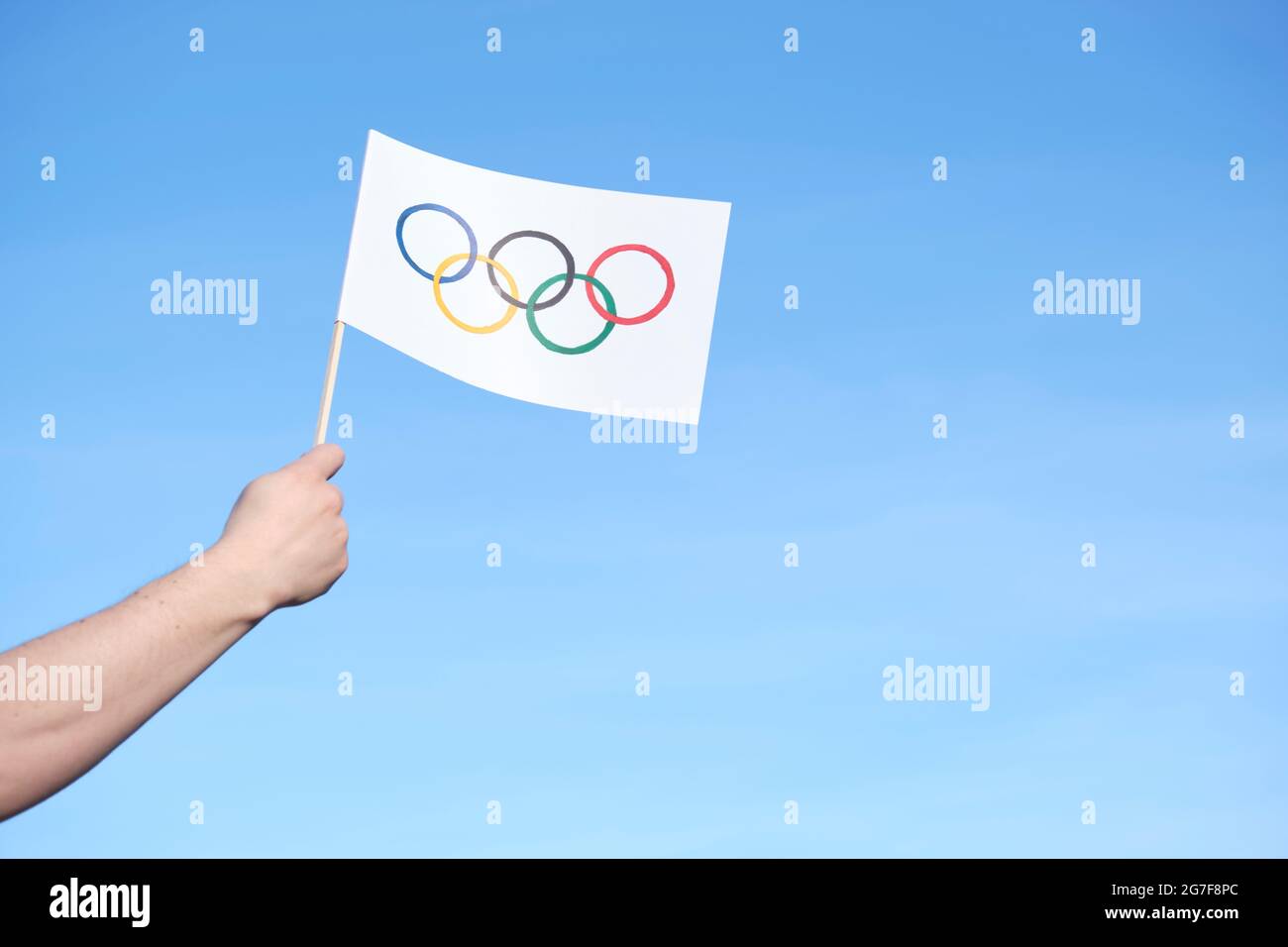 Hand holding a handmade Olympic Games flag outdoors against a clear sky on a sunny day. Image with copy space. Stock Photo