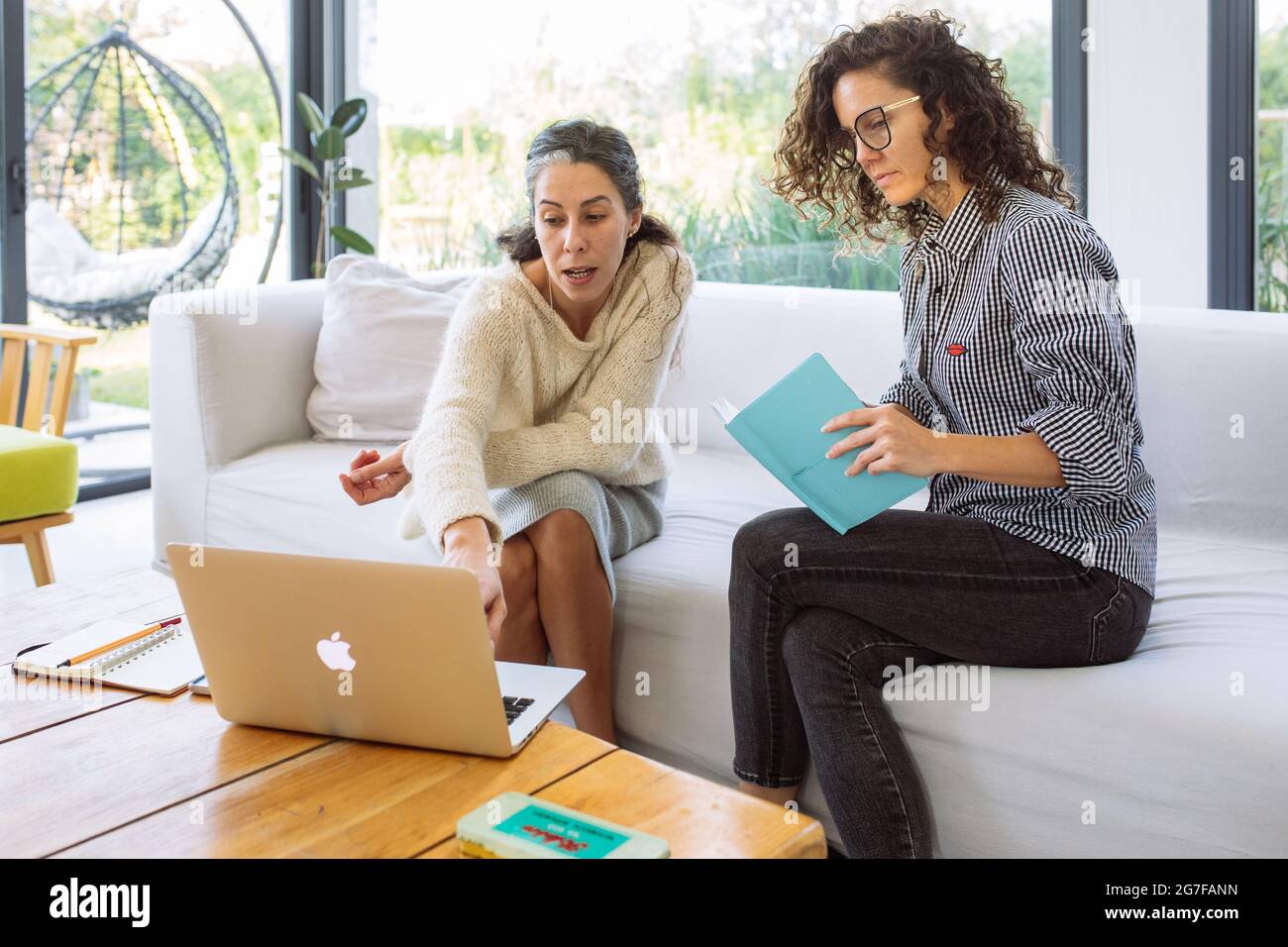 2 middle age women, a caucasian and a latino, chatting and planning sitting on a sofa in a big and bright living pointing something on a laptop Stock Photo