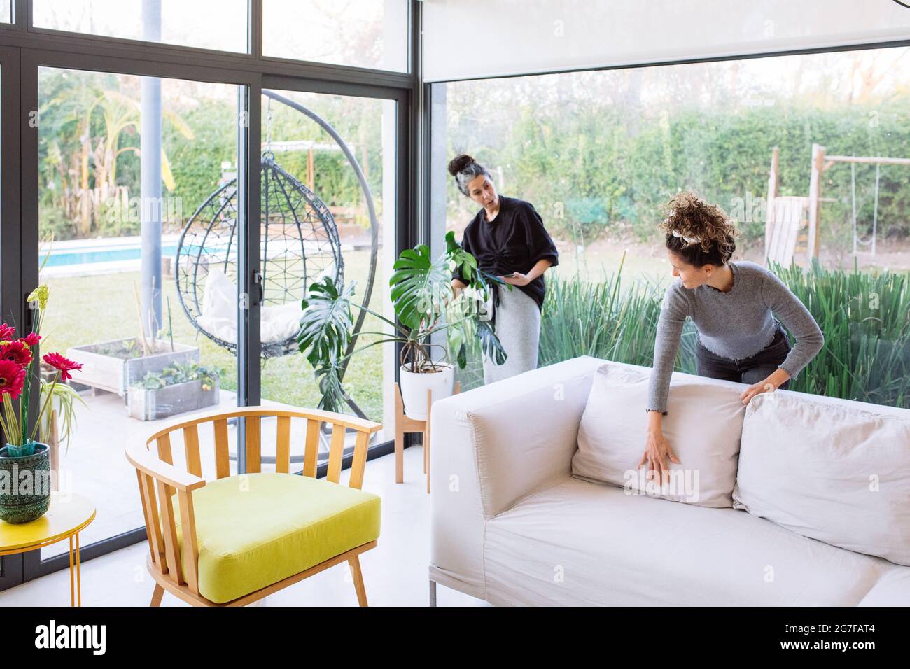 2 women, a caucasian and a latino, working on home staging a living room with big windows and a green backyard Stock Photo