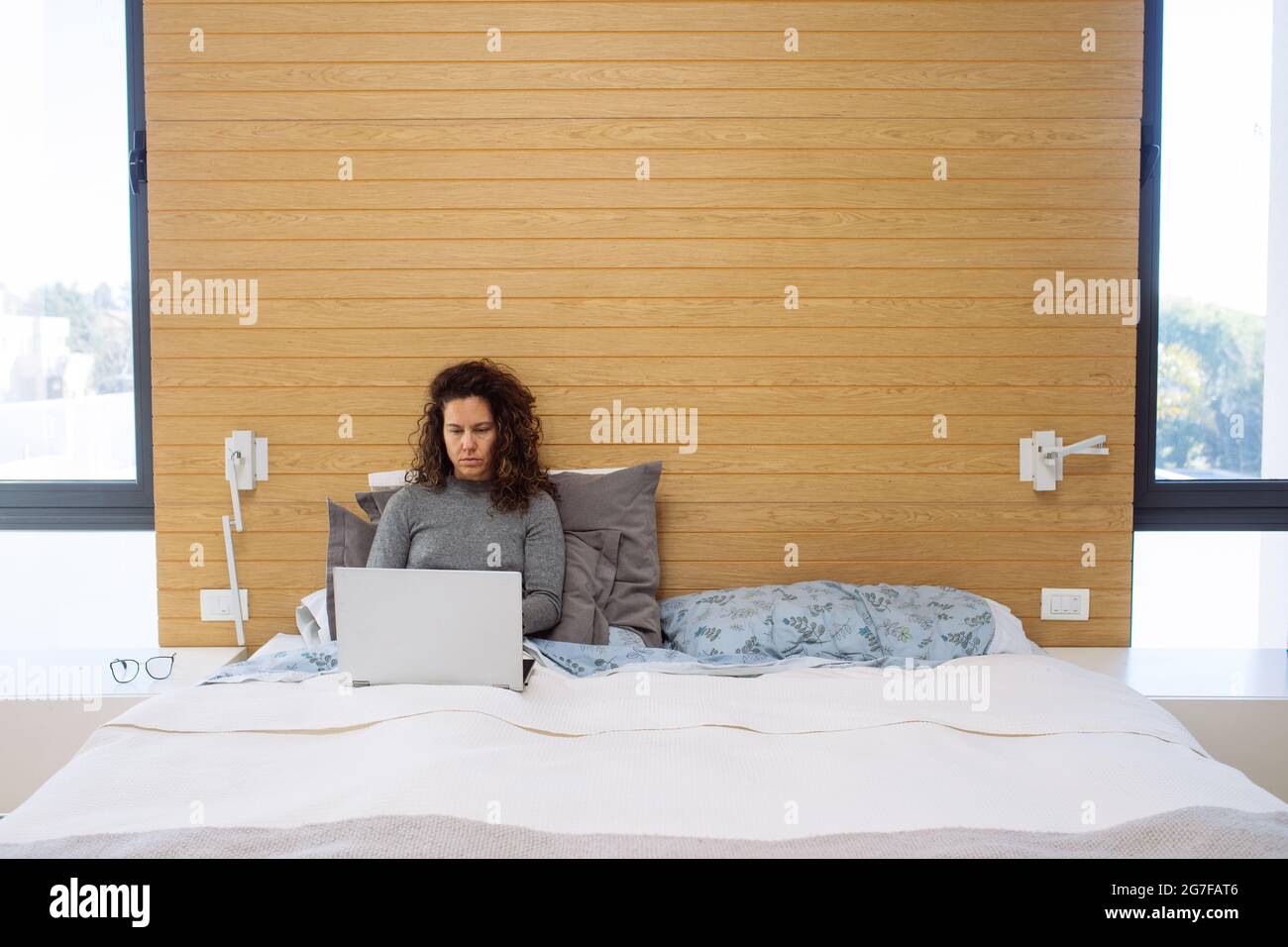 Caucasic woman in her thirties in a double king size bed working with a laptop with sleepy concentrated face Stock Photo