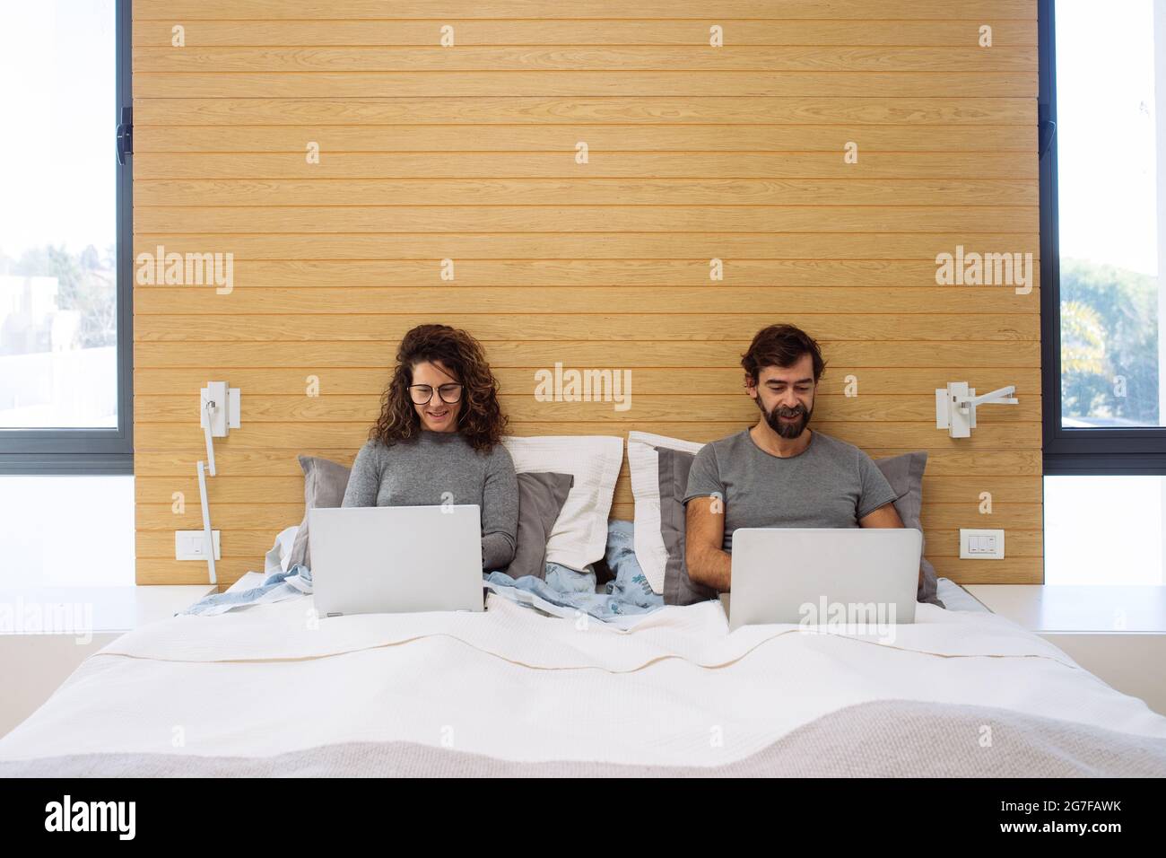 Couple in a double bed king size each with their notebook working watching each of their screens and smiling. Concept: home office working from bed Stock Photo