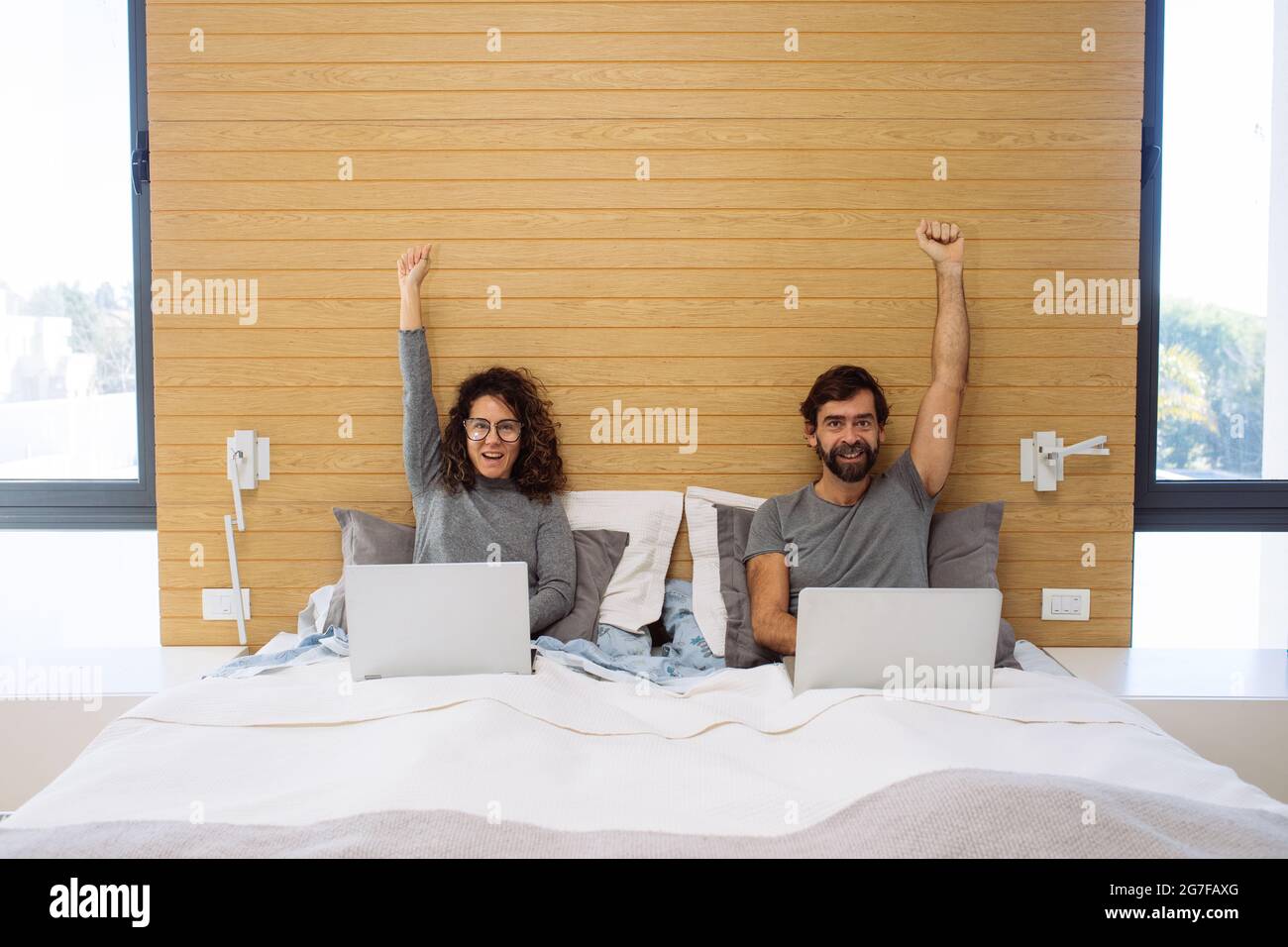 Couple in a double bed king size each with their notebook with arms up and fist closed looking at each other. Concept: home office success Stock Photo