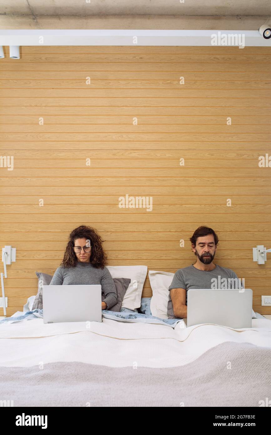 Couple in a double bed king size each with their notebook working watching each of their sreens. Concept: home office working from bed Stock Photo