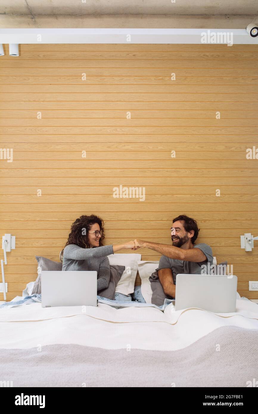 Couple in a king size double bed each with their notebook fist bumping and looking at each other. Concept: home office success Stock Photo