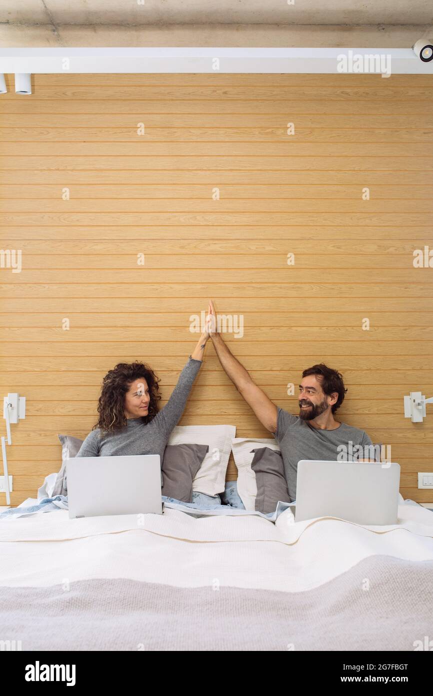 Couple in a king size double bed each with their notebook doing a hi five looking at each other. Concept: home office success Stock Photo