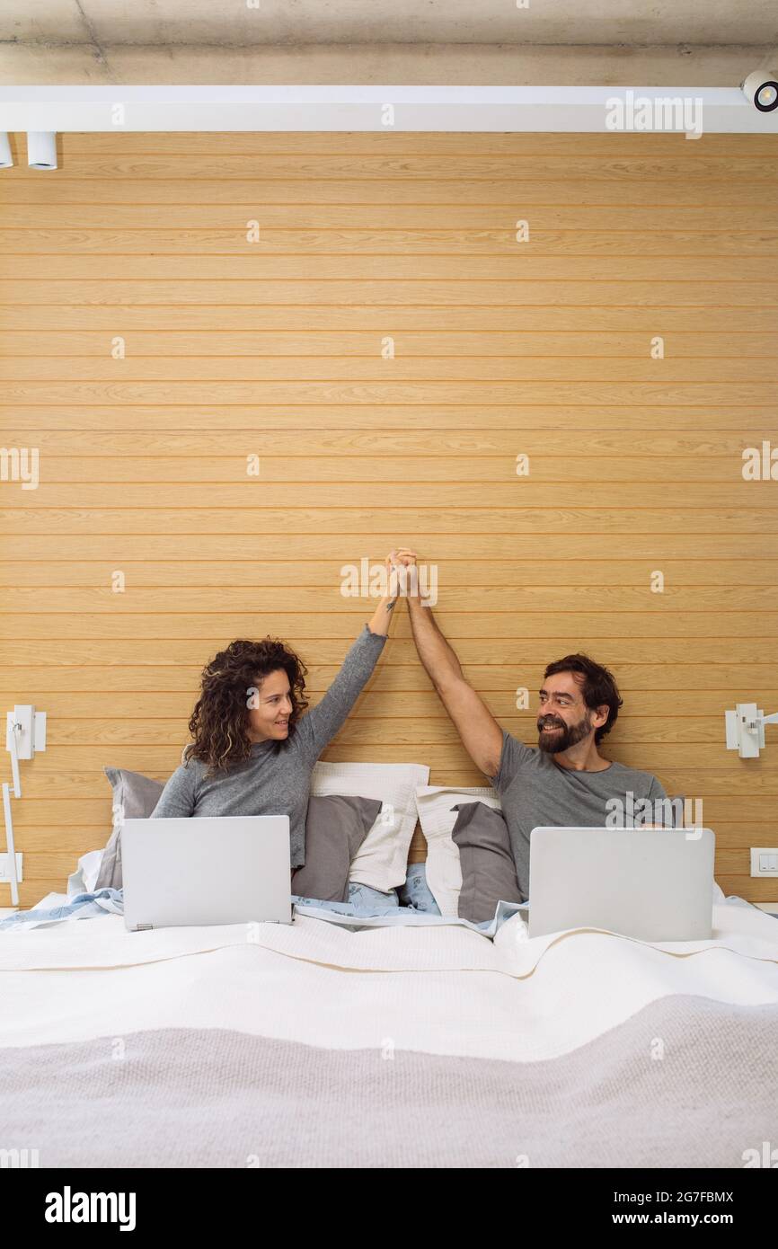 Couple in a king size double bed each with their notebook doing a hi five looking at each other. Concept: home office success Stock Photo