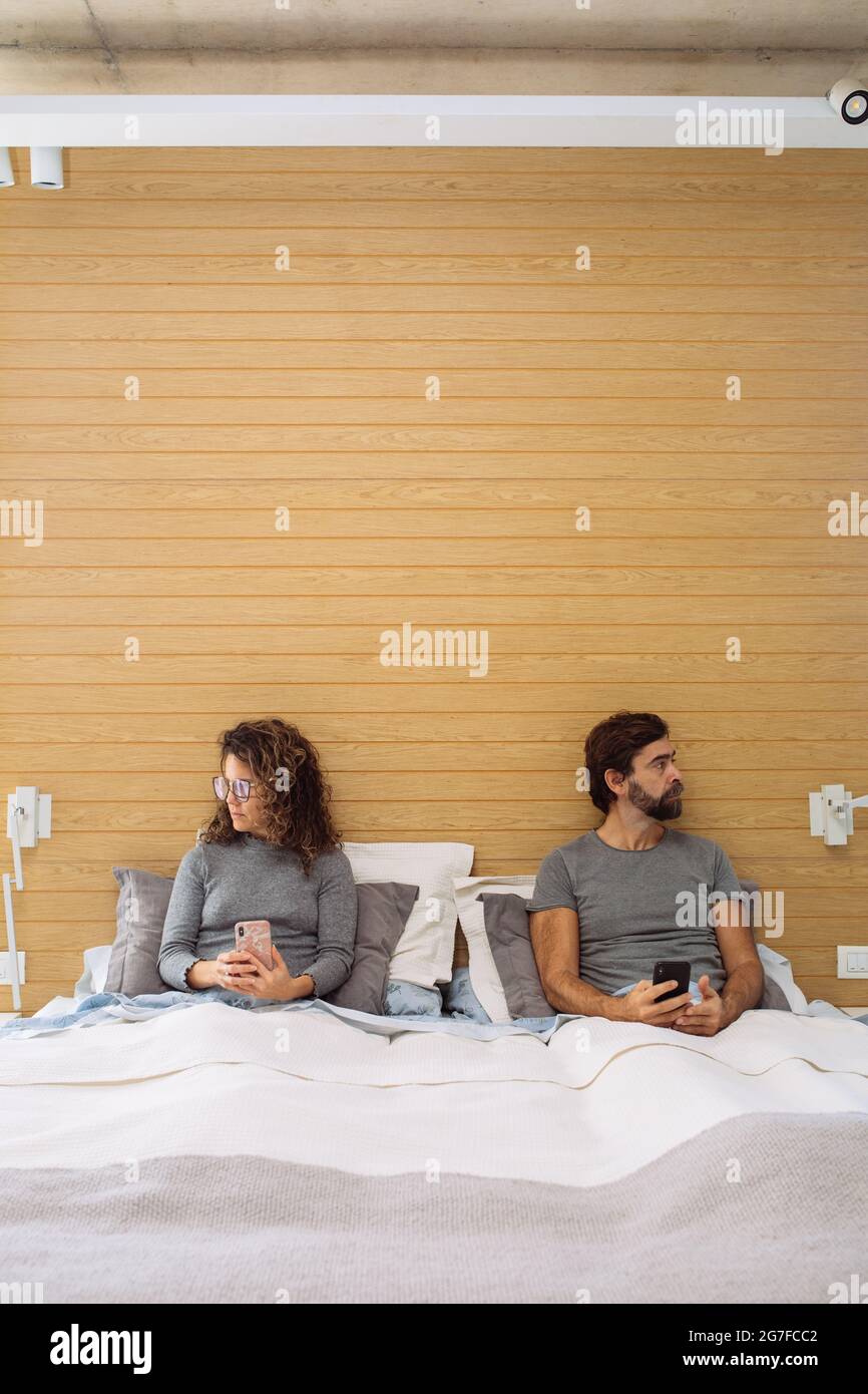 Middle age couple in a double bed king size with mobile phone in their hand looking at opposite directions. Concept: disconnection Stock Photo