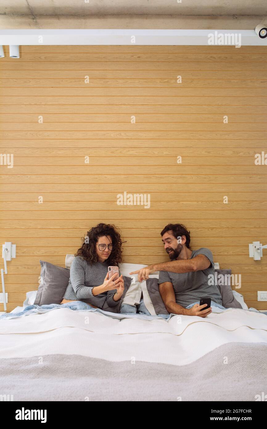 Middle age couple in a double bed king size where the woman is showing the man something on his mobile phone and the man is pointing at the screen Stock Photo