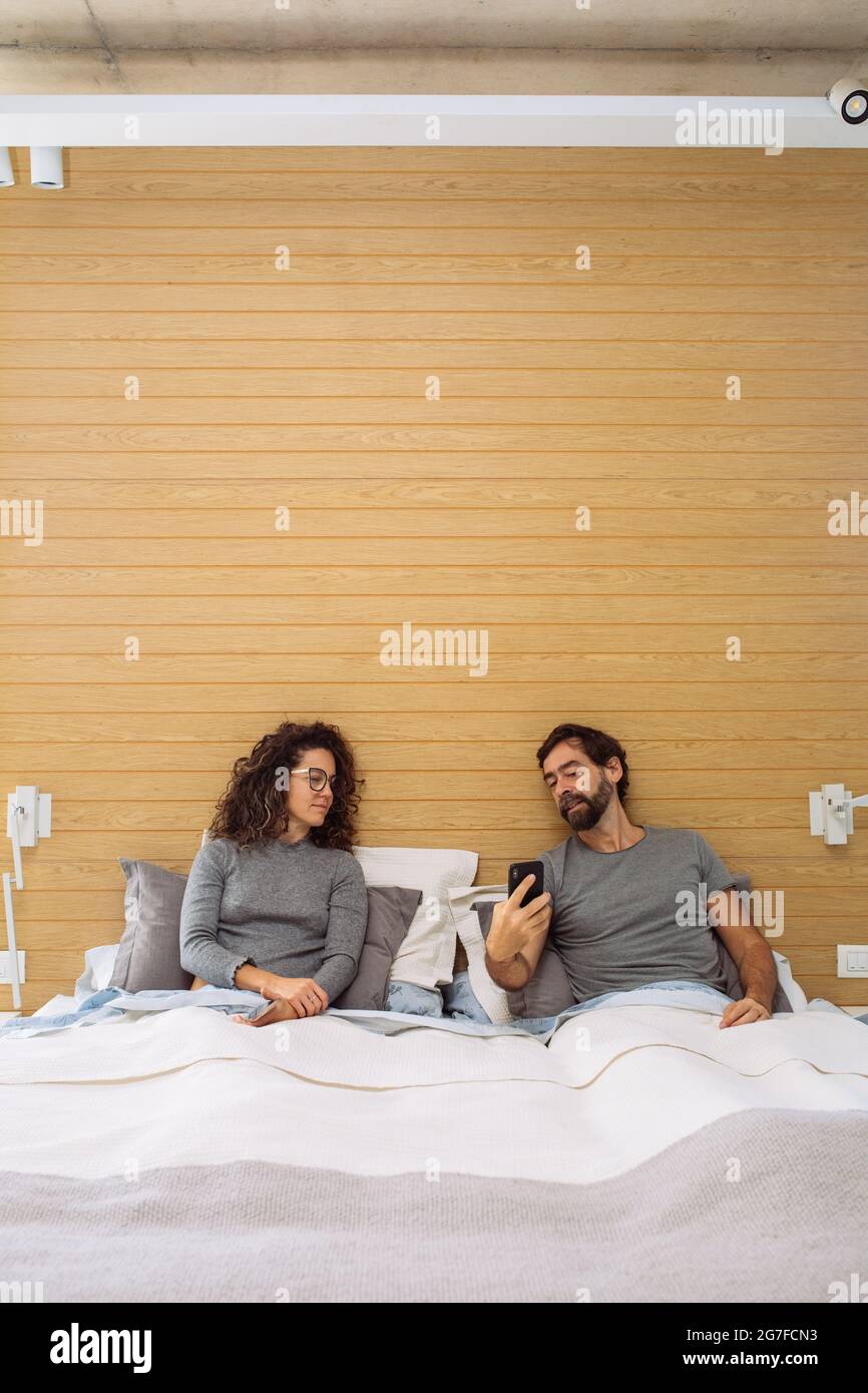 Middle age couple in a double bed king size where the man is showing the woman something on his mobile phone screen Stock Photo