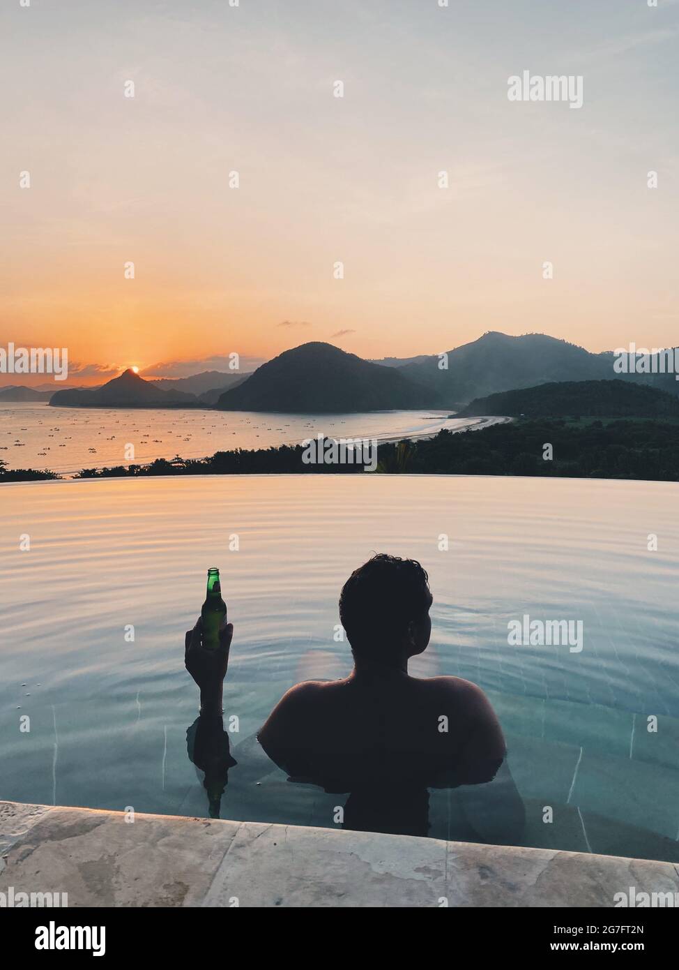 Silhouette of a man with a beer swimming in luxury infinity pool during ...