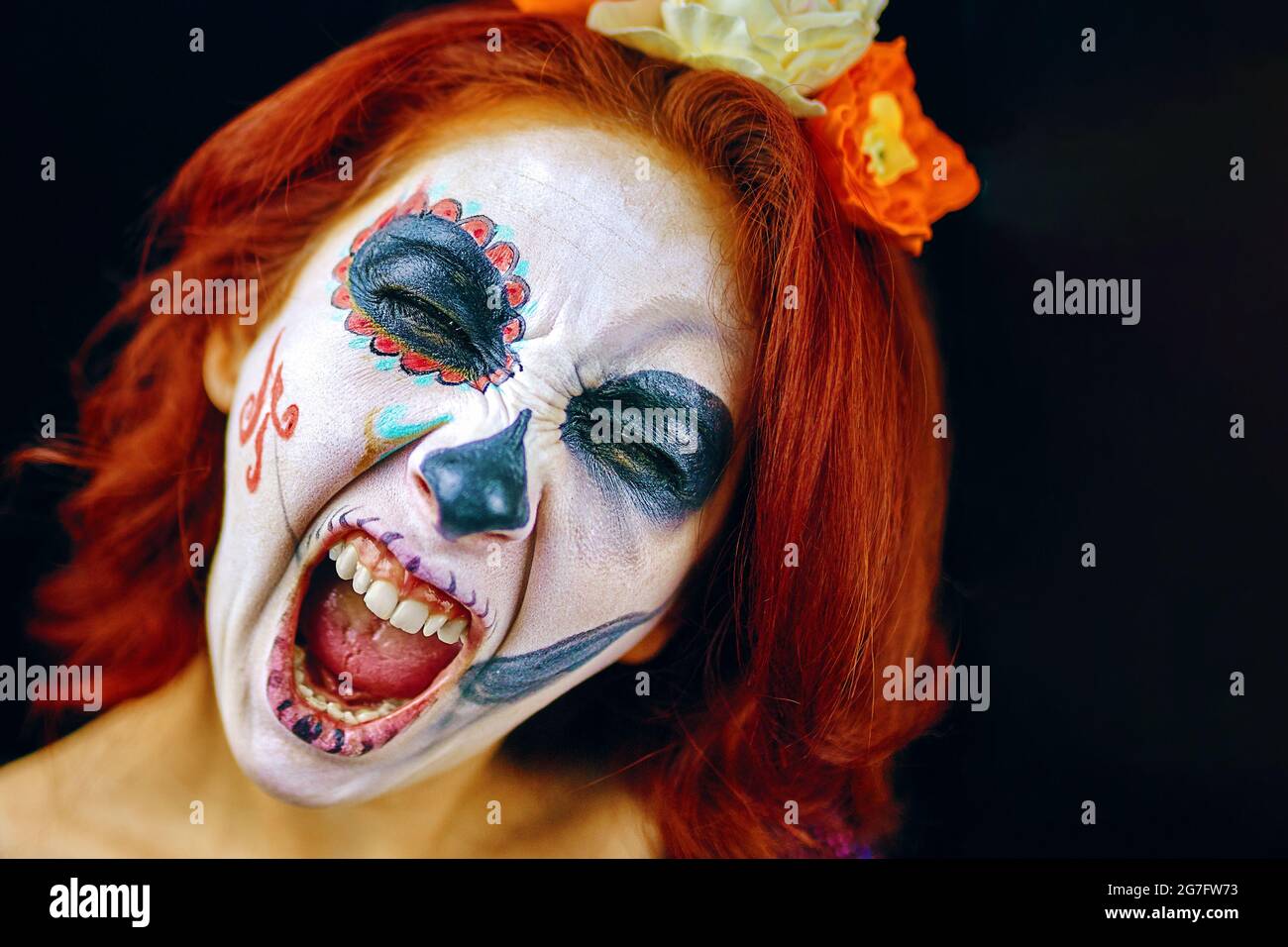 A young woman in day of the dead mask skull face art. Woman with skull makeup and red hair, screaming on dark background close up Stock Photo