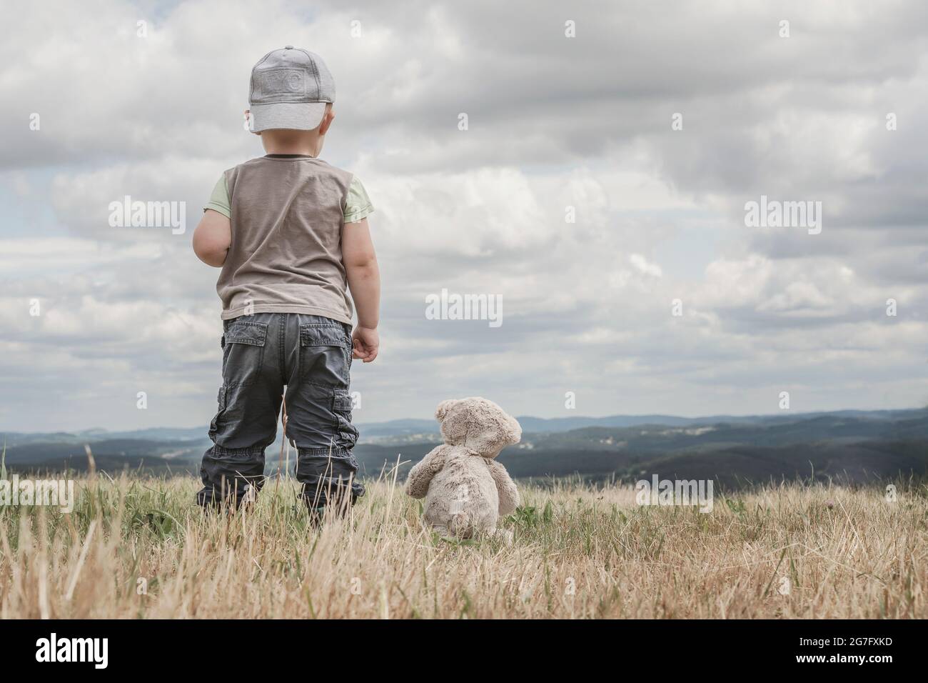 Two Year Old Boy In Cap And Pocket Pants Stand On Top Of Hill Back To Camera With Plush Teddy Bear Side By Side Stock Photo Alamy