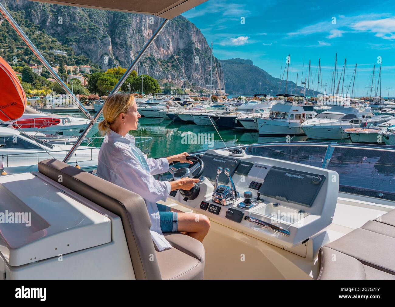 Beautiful blond woman driving yacht. Adult woman model in a white shirt sitting at the wheel of the yacht and begin to drive luxury boat from the port. Stock Photo