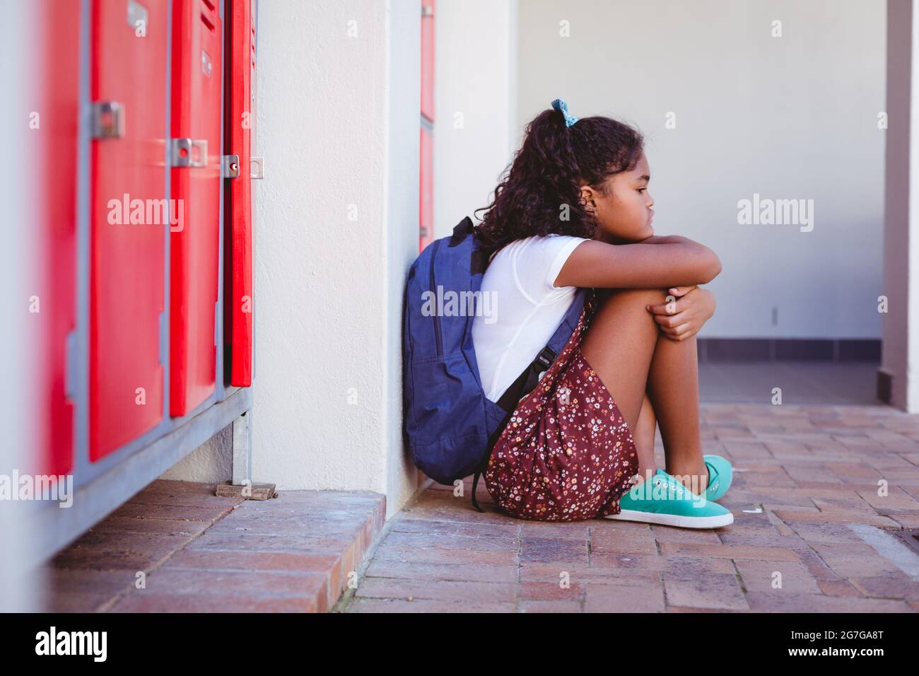 Unhappy african american schoolgirl sitting by lockers in school corridor with schoolbag Stock Photo