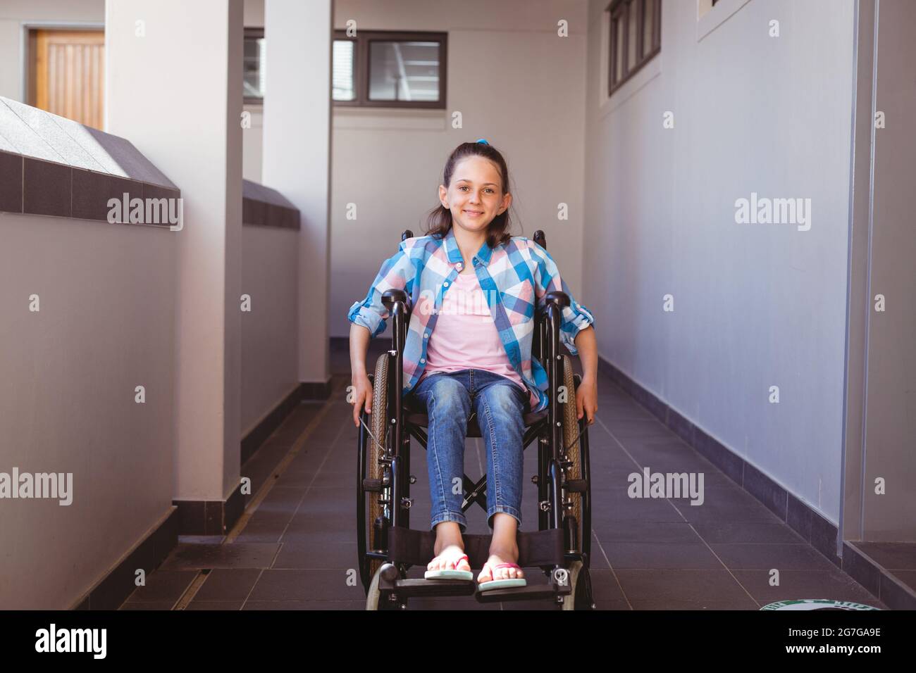 Portrait of smiling disabled caucasian schoolgirl sitting in wheelchair in school corridor Stock Photo