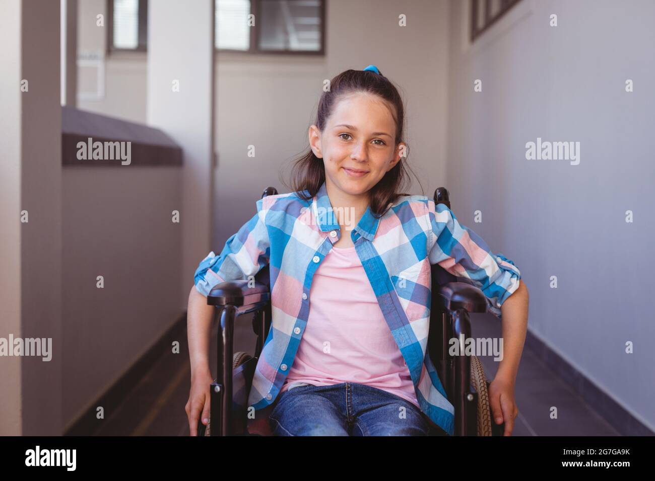 Portrait of smiling disabled caucasian schoolgirl sitting in wheelchair in school corridor Stock Photo