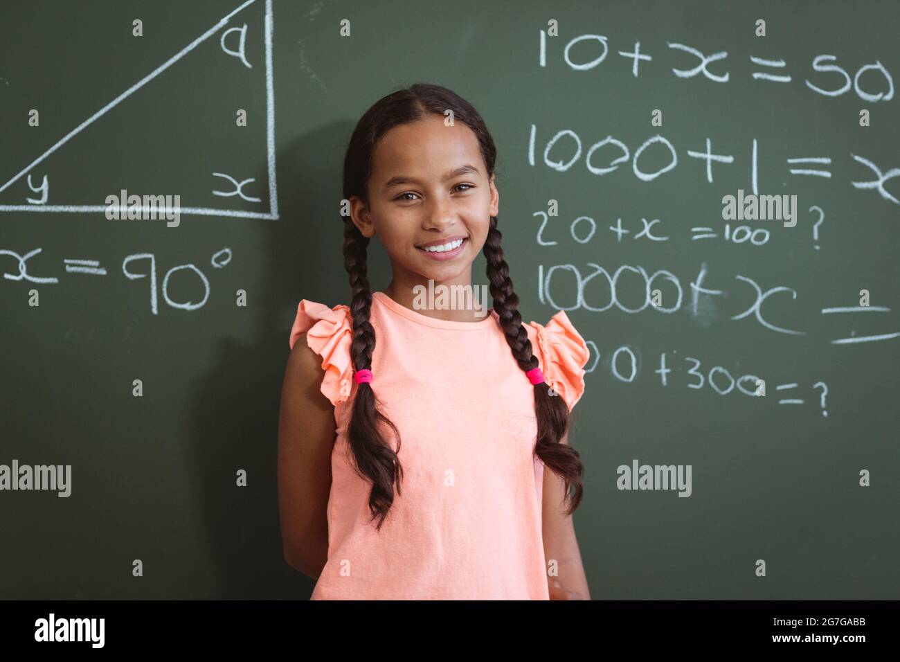 Portrait of smiling mixed race schoolgirl standing in front of chalkboard in classroom maths lesson Stock Photo
