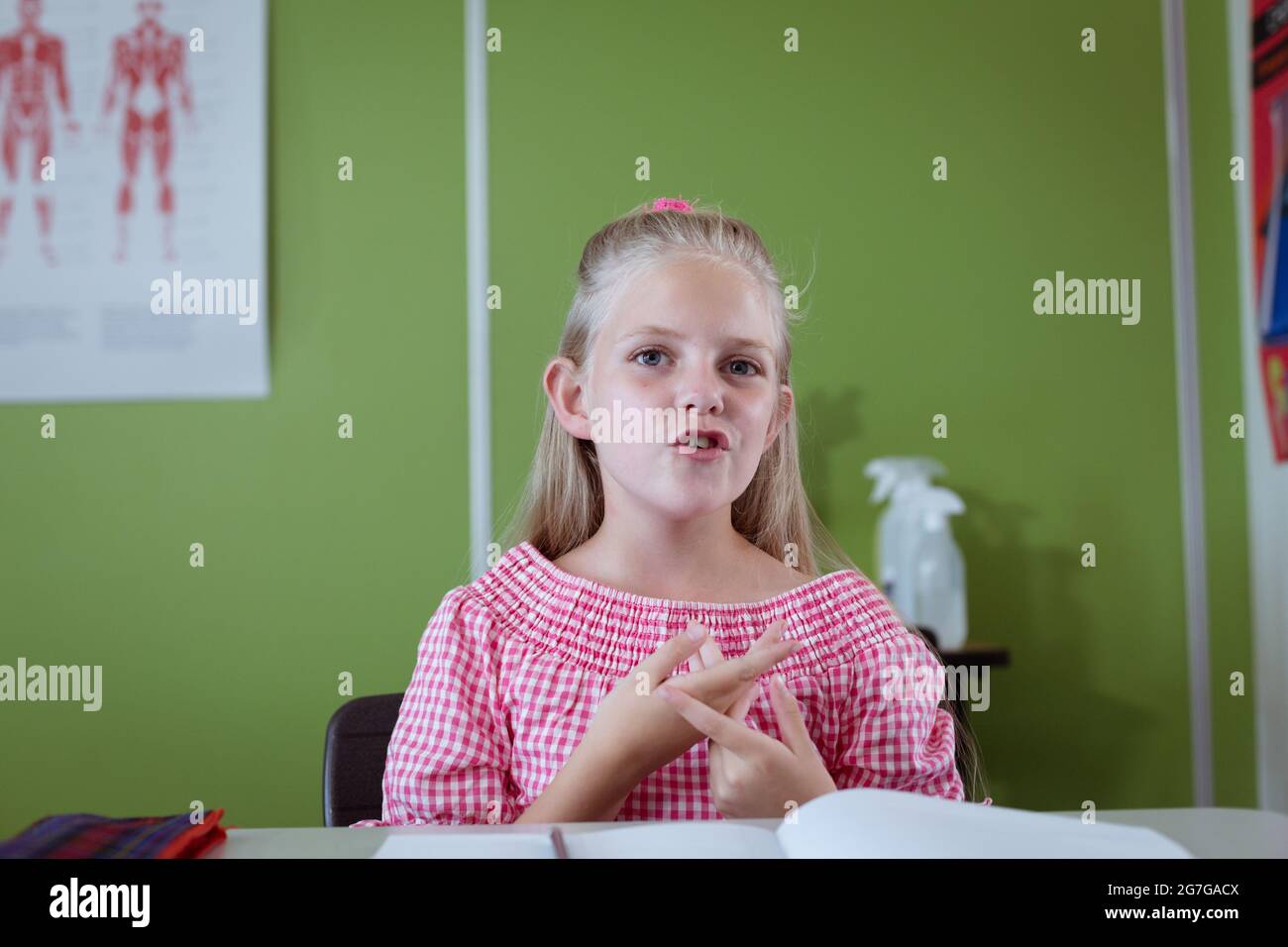 Portrait of caucasian schoolgirl sitting at desk in classroom talking ...