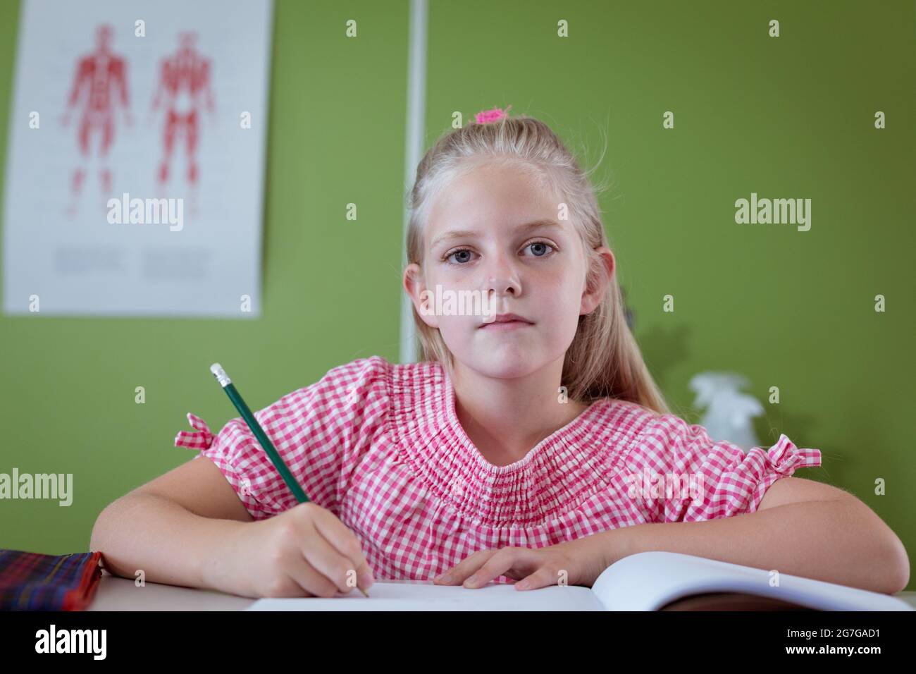 Portrait of caucasian schoolgirl sitting at desk writing in classroom ...