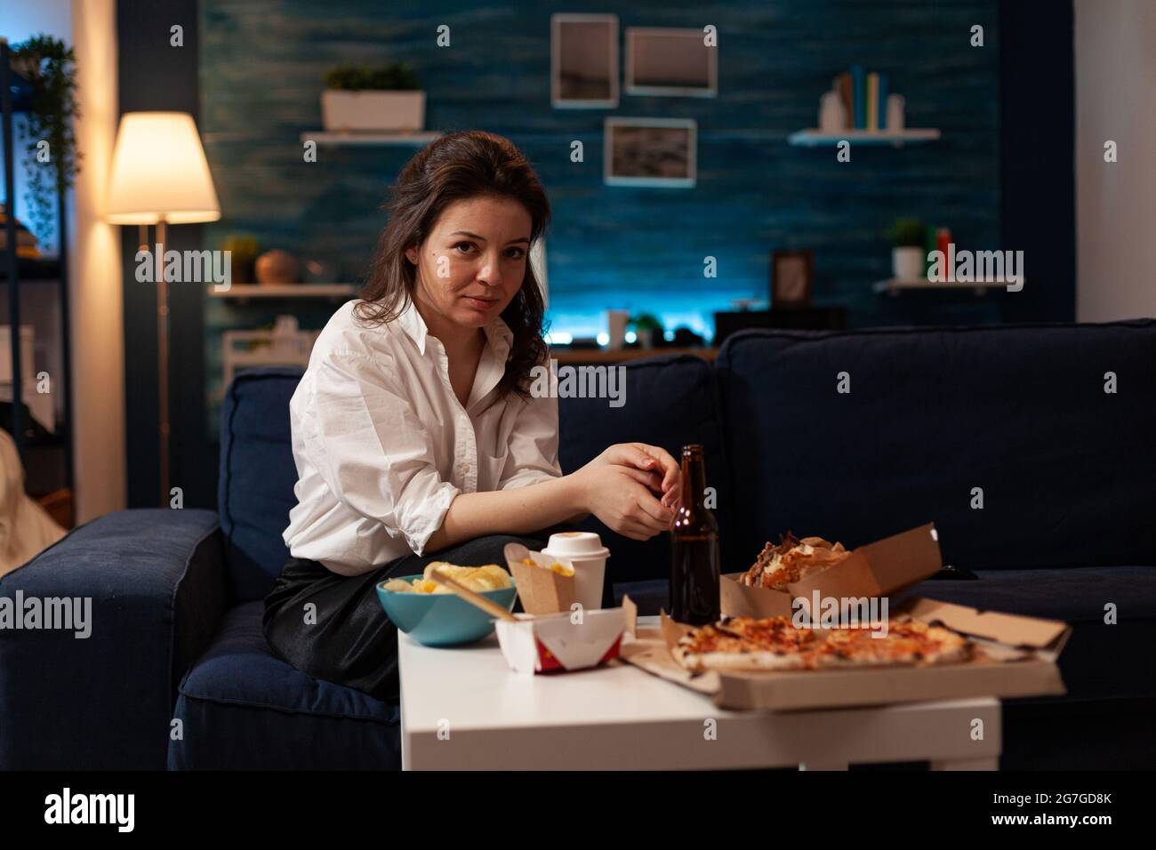 Portrait of smiling woman looking into camera sitting on couch enjoying free time ordering takeaway meal in living room at night. Junk food standing on table. Fast food order. Delivery fast-food box Stock Photo