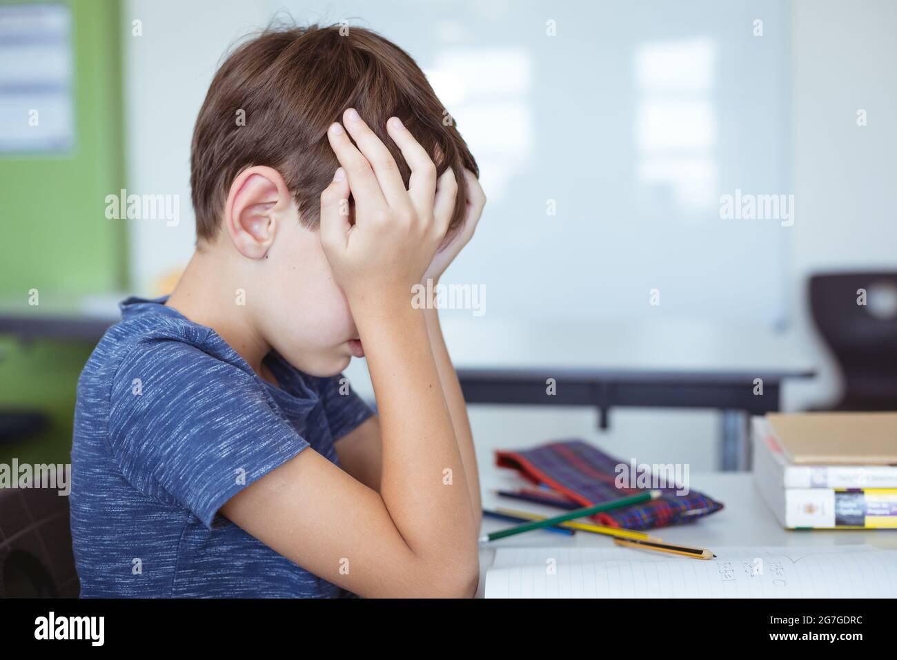 Tired caucasian schoolboy in classroom sitting at desk rubbing eyes Stock Photo