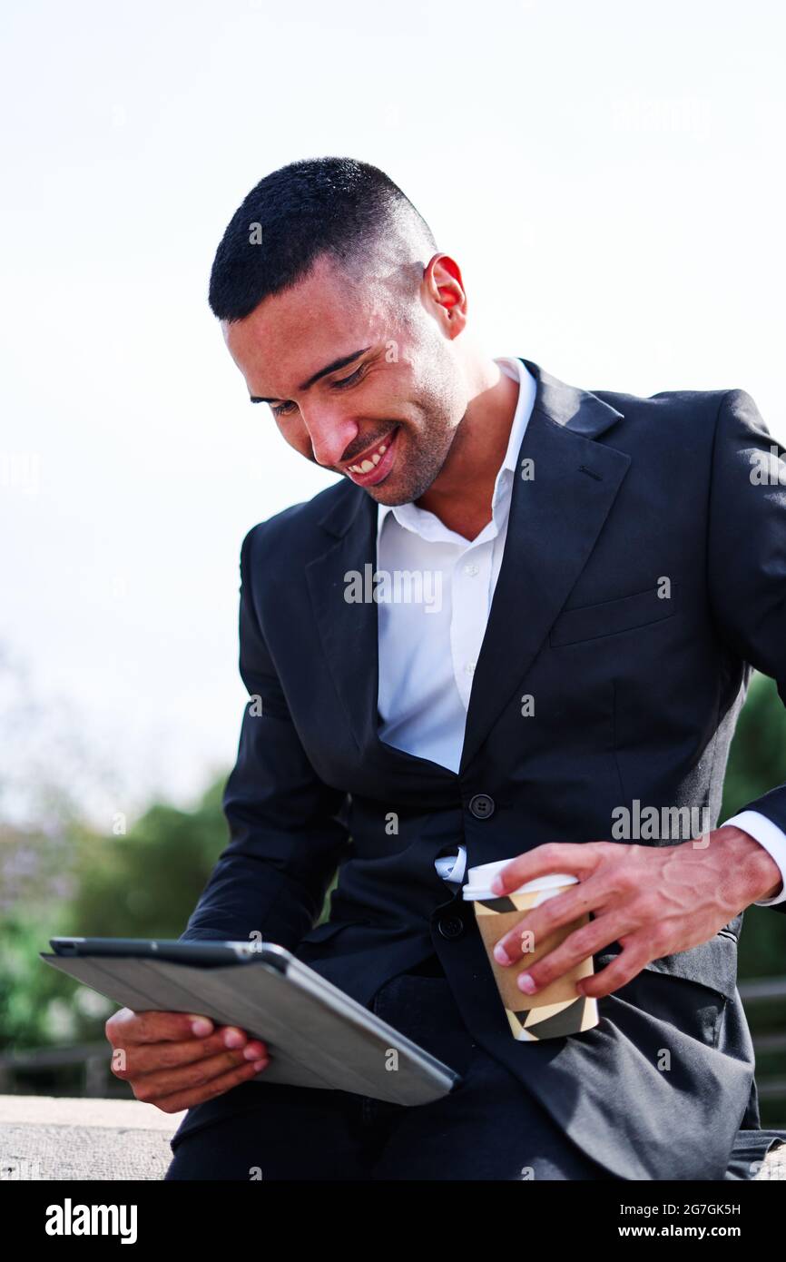 Side view of happy young Hispanic man in elegant formal suit drinking takeaway beverage and browsing tablet while having free time and resting in park Stock Photo