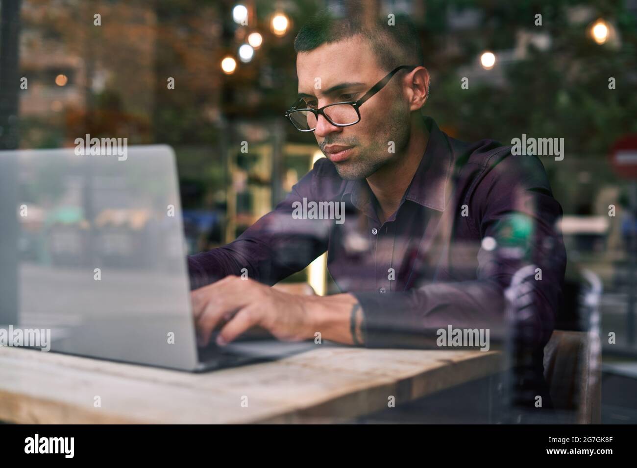 Serious Hispanic male manager in violet shirt and glasses typing on netbook keyboard while sitting at table behind window and working on project in ca Stock Photo