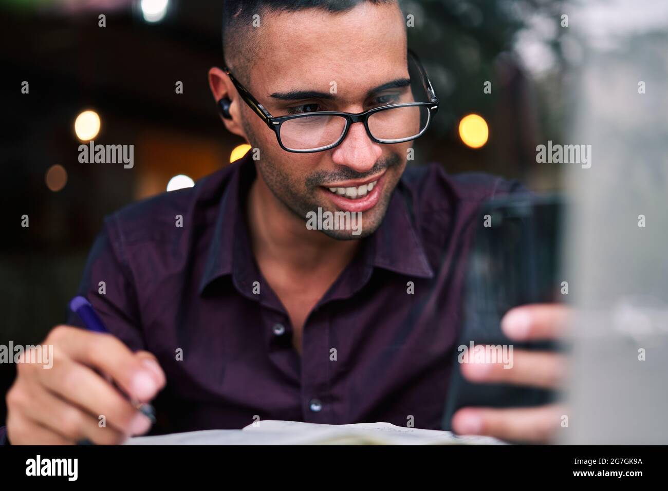 Handsome young Hispanic male entrepreneur checking information on smartphone and writing notes in planner while working at table with laptop Stock Photo