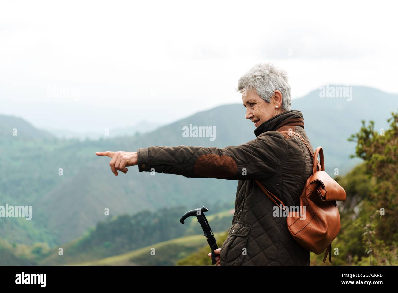 Side view of elderly Woman with backpack and trekking stick standing on grassy slope towards mountain peak during trip in nature pointing with finger Stock Photo