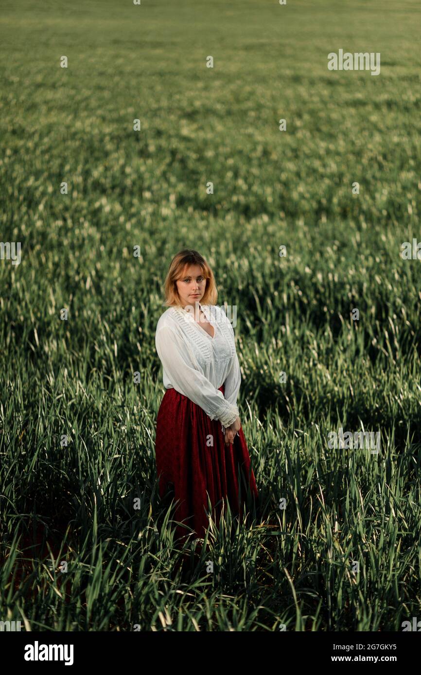 Calm young female dressed in old fashioned blouse and skirt standing alone among tall green grass in cloudy summer day in countryside Stock Photo