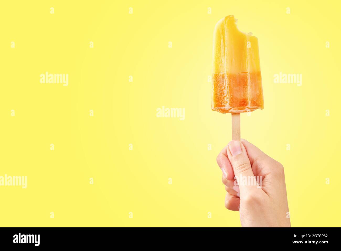 Woman holds bitten ice cream on yellow background with copy space. Color frozen fruit popsicle. Stock Photo