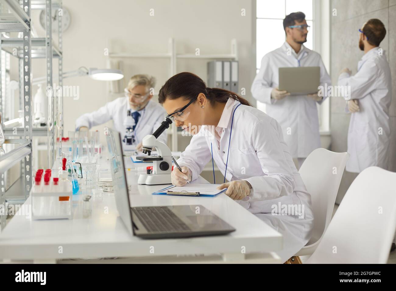 Young woman lab worker working with microscope making notes on clipboard Stock Photo