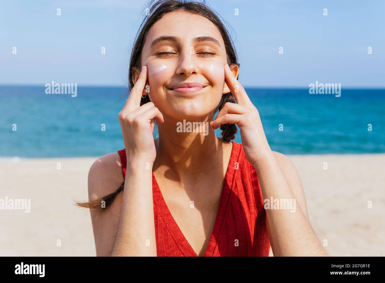 Smiling female with closed eyes applying suntan lotion on face on sunny