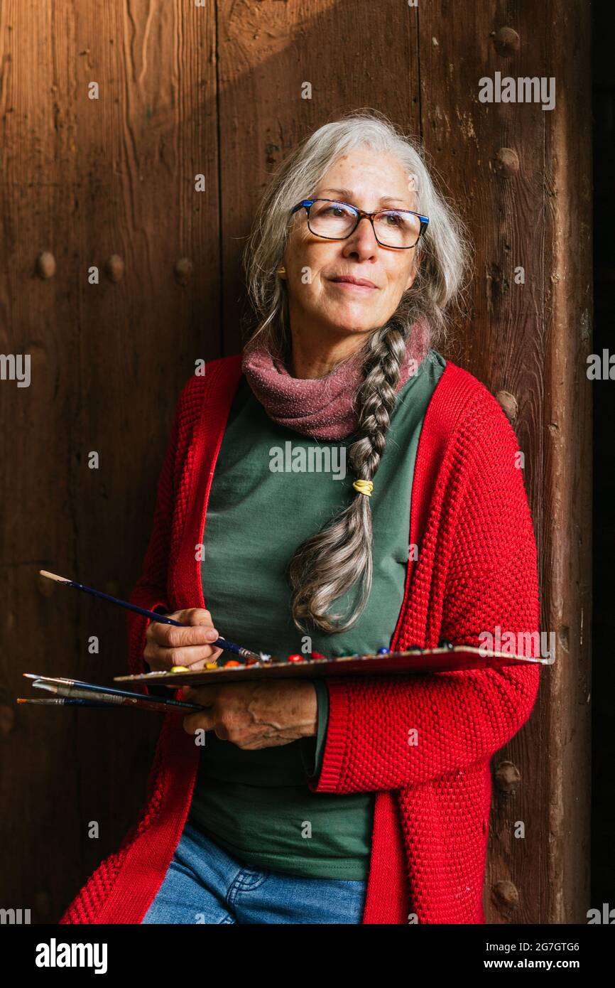 Delighted senior female artist with paint palette and brushes standing ...