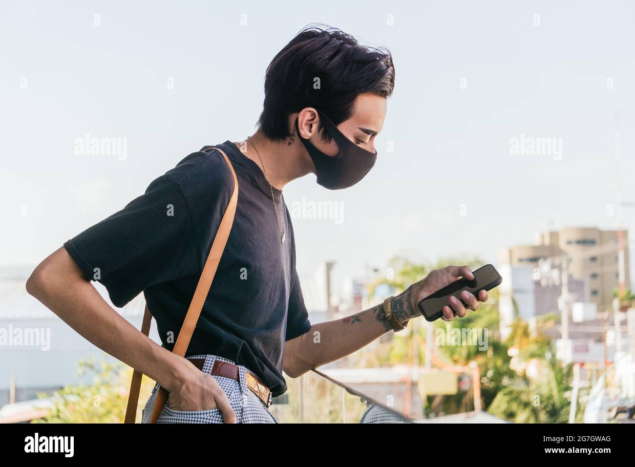 Side view of homosexual man in protective mask using mobile phone while standing on balcony in city during coronavirus Stock Photo
