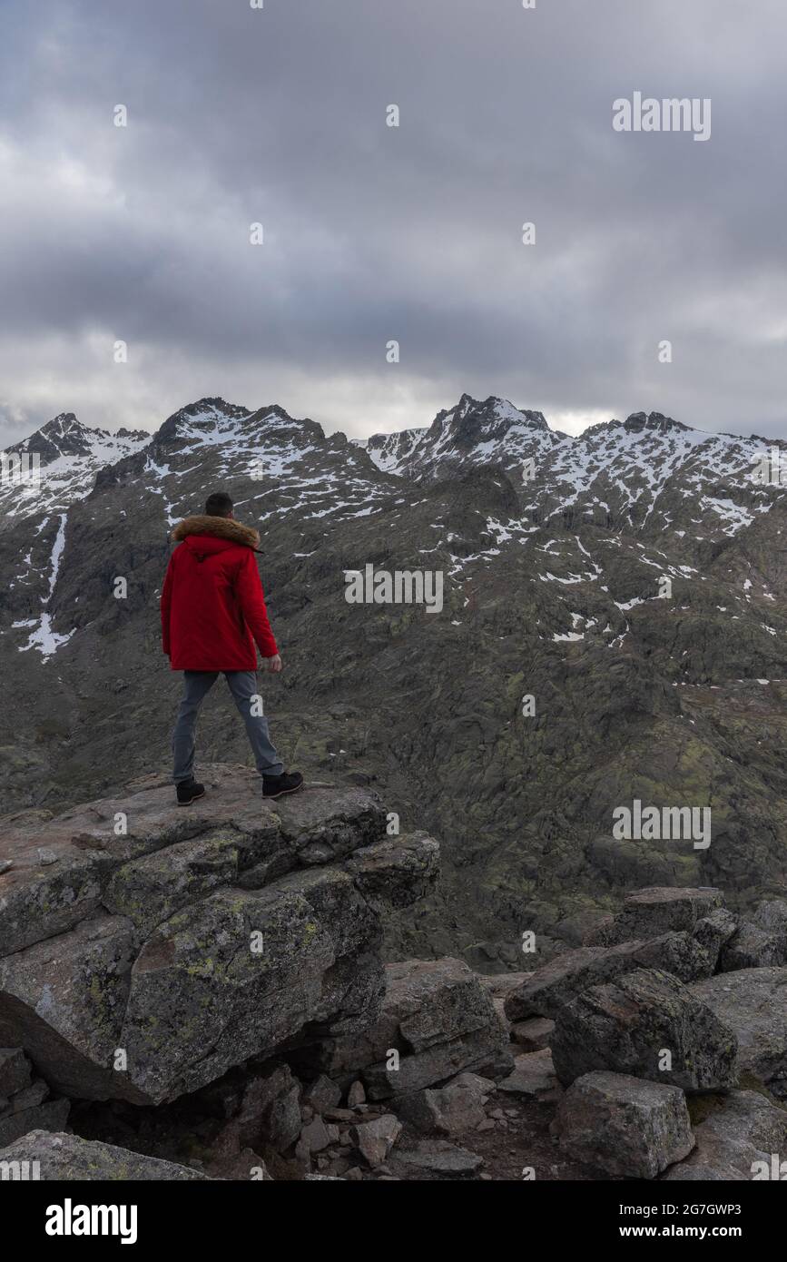 Back view of unrecognizable man in outerwear standing on stone and looking at snowy Sierra de Gredos mountain ridge in cloudy evening in Avila, Spain Stock Photo