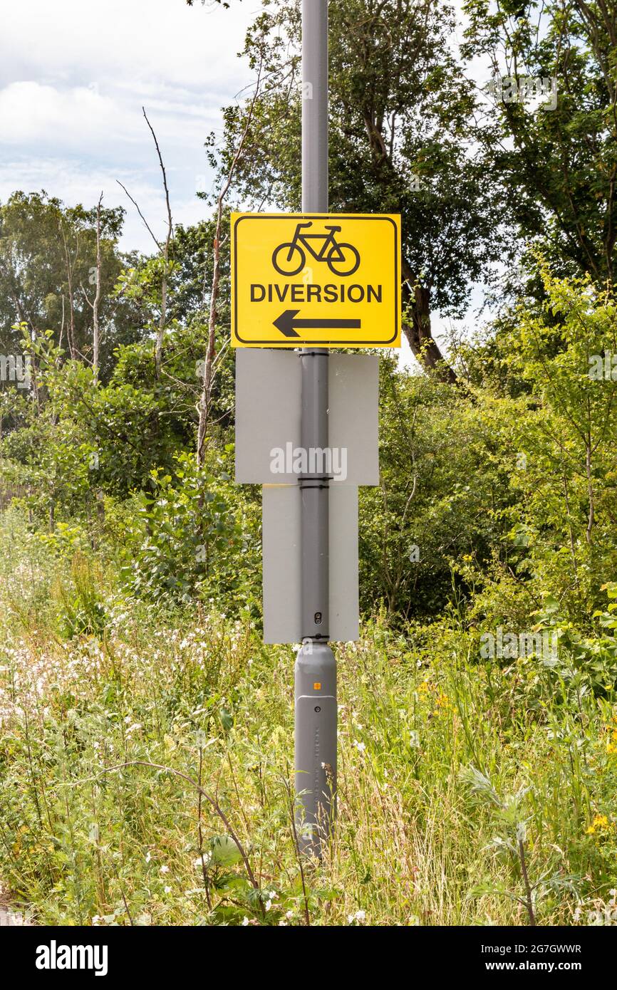 A yellow bicycle path diversion sign on a lampost on a overgrown verge in Cambridge, UK. Stock Photo