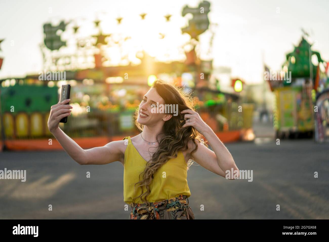 Content female taking self portrait on mobile phone while having fun in amusement park in evening in summer Stock Photo