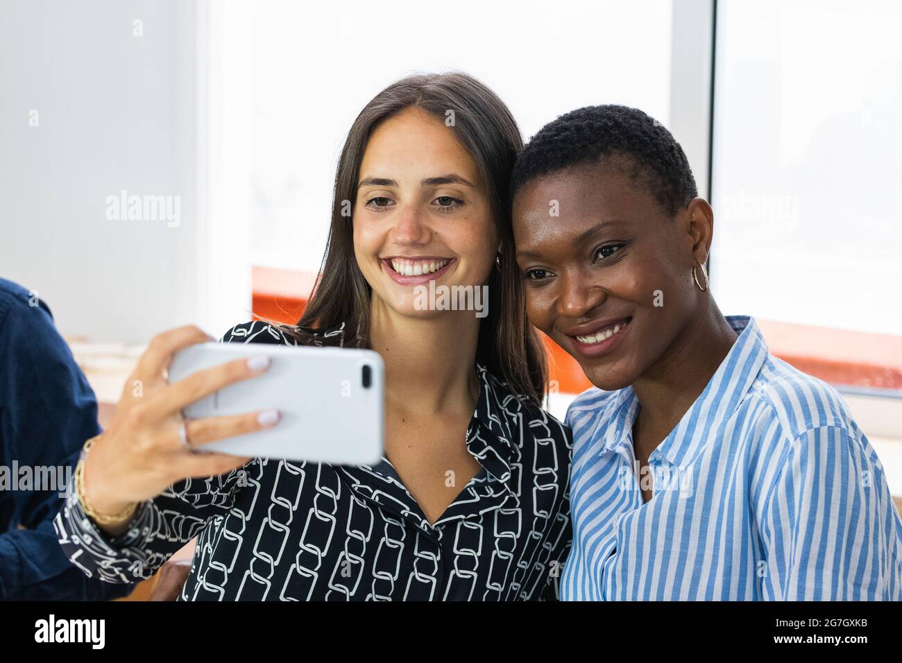 Cheerful multiracial female colleagues taking self portrait on smartphone while having lunch together in kitchen at workplace Stock Photo