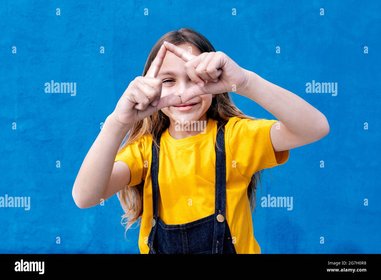 Cheerful teenager looking through hole of triangle hand sign on blue background in studio Stock Photo
