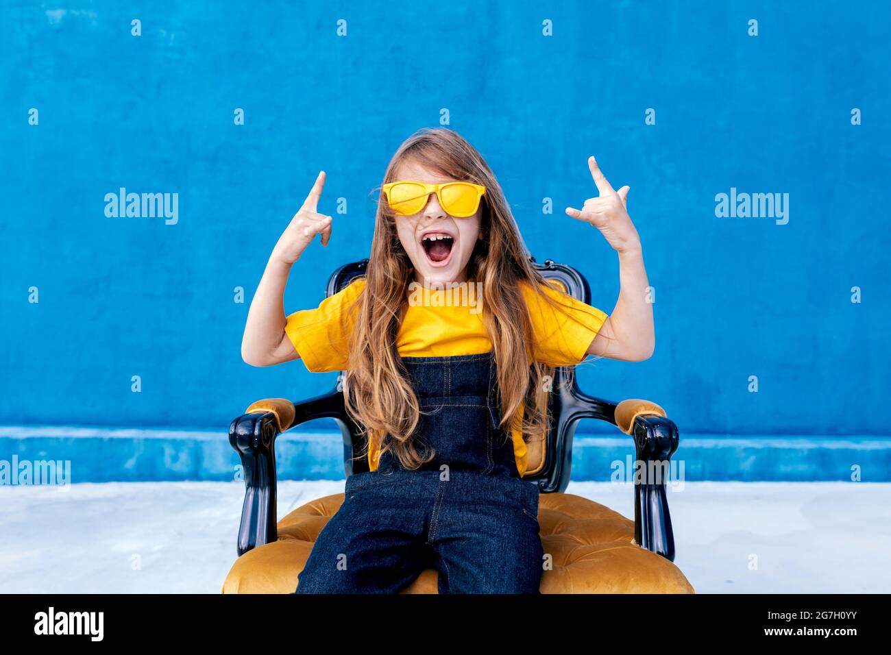 Expressive teenager with long hair and in trendy yellow sunglasses showing rock sign and screaming on blue background Stock Photo