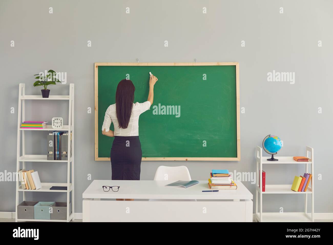 Back view of a young school teacher writing on the green chalkboard in the classroom Stock Photo