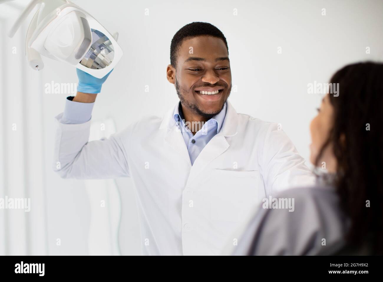 Handsome Black Stomatologist Doctor Ready For Check Up With Female Patient, Turning On Lamp Before Starting Treatment. Dentist Man Looking At Customer Stock Photo