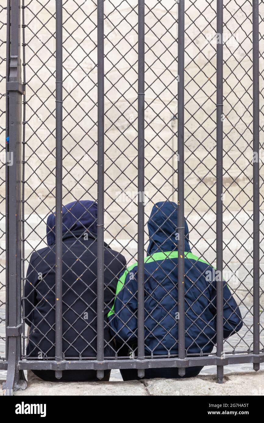 Two people seated behind a fence and a wire mesh, hooded, seen from behind. Paris, France Stock Photo