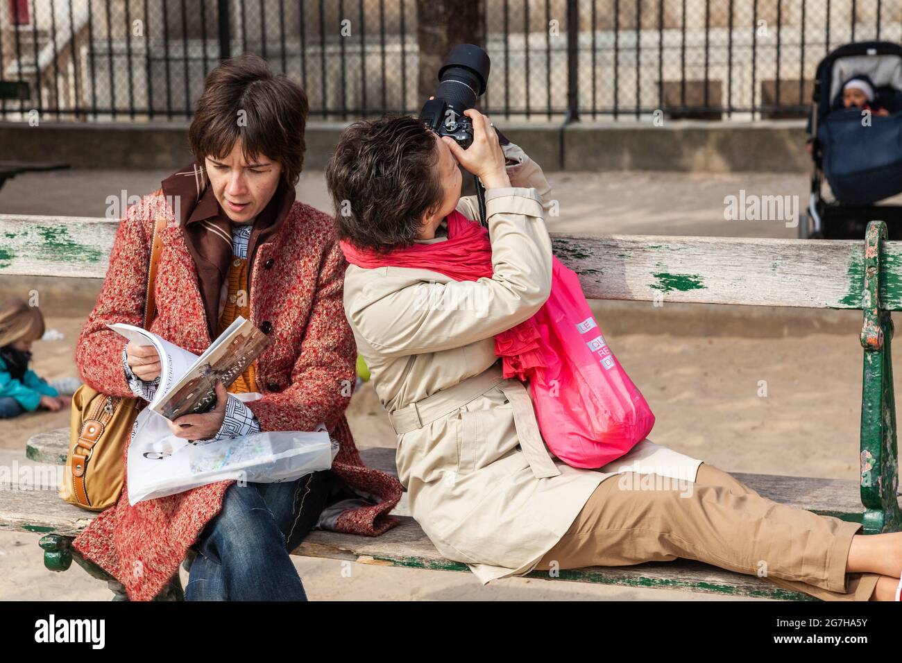 Two ladies seated on a bench, one leafing through a book, the other photographing. Paris, France Stock Photo