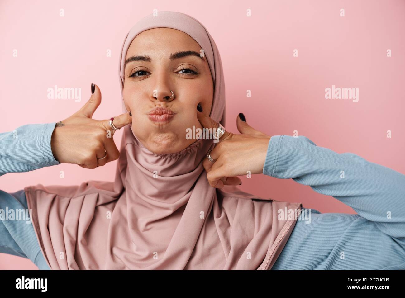 A portrait of the positive muslim woman wearing pink hijab puffed out cheeks while standing in the pink studio Stock Photo