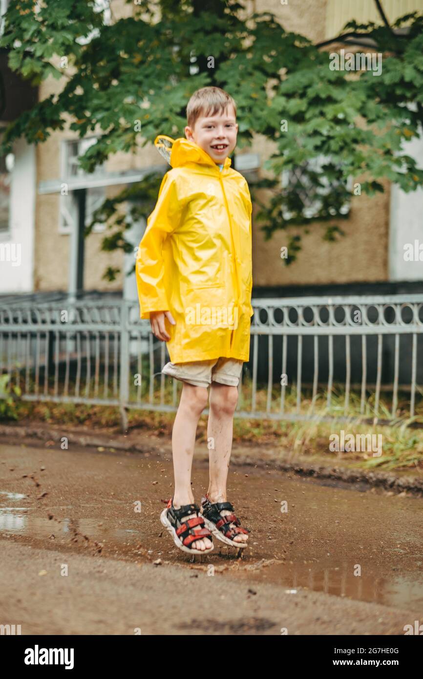 A boy in a yellow raincoat walks outside in the rain. A child jumps on the narrows. jump through muddy puddles. Happy and carefree childhood Stock Photo Alamy